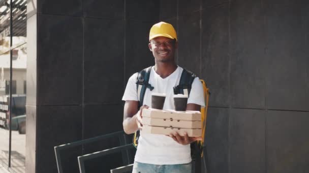 Молодий африканський чоловік доставки в білій формі з коробками для піци, обідньою коробкою і кавою, дивлячись на камеру і посмішку, готовий обслуговувати клієнтів. Служба доставки, замовлення на винос. Крупним планом коробка для піци — стокове відео