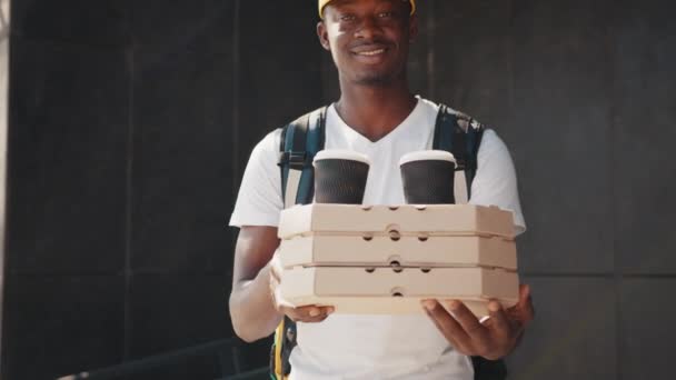 Pizzásdoboz bezárva. Fiatal afrikai futár fehér egyenruhában pizzás dobozokkal, uzsonnás dobozzal és kávéval, kamerába néz és mosolyog, készen áll kiszolgálni ügyfeleit. Szállítási szolgáltatás, elvitelre szóló rendelések — Stock videók
