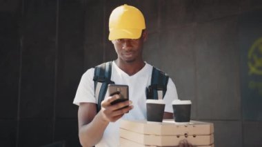 Afrikalı kurye pizza ve kahve kutusunu taşırken telefonunu kontrol ediyor. Güzel, siyah, karton kutuları olan bir erkek kurye. Modern evin dışında dururken müşteriye pizza getiriyor.