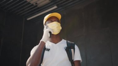 Maskeli ve eldivenli genç bir siyah kurye sipariş alan bir müşteriyle telefonda konuşuyor. Şapkalı ve koruyucu maskeli bir afro kuryenin portresi bir restorandan veya dükkandan gelen yemekleri yüceltiyor.