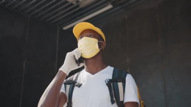 Afro kurye portresi şapka ve koruyucu maske takmış bir restorandan ya da dükkandan gelen yemekleri övüyordu. Maskeli ve eldivenli genç bir siyah kurye sipariş alan bir müşteriyle telefonda konuşuyor.
