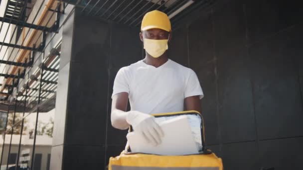 Trabajador de reparto afroamericano en gorra amarilla y camiseta blanca, máscara facial uniforme, sosteniendo caja de cartón vacía. Entrega en línea de mercancías en cuarentena. Entrega en tienda online — Vídeos de Stock