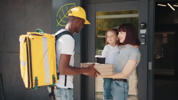 Un corriere afro-americano con una borsa termica consegna cibo e bevande a una bella ragazza con un bambino. Il giovane corriere consegna il pacco direttamente a casa dei clienti. Concetto corriere — Video Stock