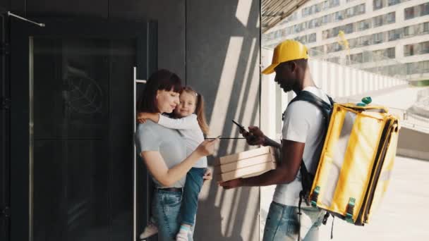 Афро-американский продавец с коробками горячей пиццы. Молодая улыбающаяся женщина с девушкой у двери, оплатить по карте для онлайн-заказа. Быстрая доставка домой. Семья: мать, дочь — стоковое видео