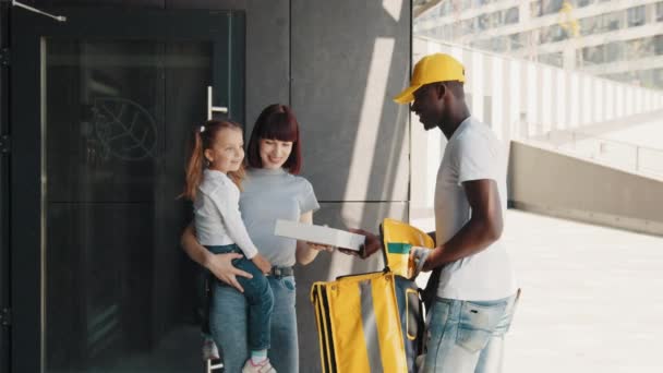 En ung kvinna med en liten flicka möter en afroamerikansk kurir. En afrikansk amerikansk kurir ger en liten flicka ett paket eller en gåva från en webbutik. Kuriren lämnar lådan till barnet. — Stockvideo