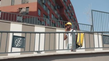 Termal sırt çantalı bir Afro-Amerikan kurye bir müşteriyle telefonda konuşuyor, caddede yürüyor, müşterilere siparişleri iletiyor. Şık şehir binaları olan modern bir şehirde güneşli bir gün.