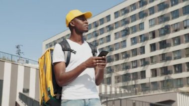 Yakışıklı Afro-Amerikan yemek dağıtım kuryesi kameraların önünde sokakta poz veriyor. Sarı şapkalı gülümseyen bir adam ve sırtında ısı yalıtımlı bir yemek torbası elinde bir telefonla.