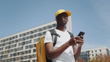 Bir Afro-Amerikalı kurye termal sırt çantasıyla yiyecek getiriyor, sokakta duruyor, akıllı telefondan navigasyona bakıyor. Tedarikçinin çalışanı siparişi çevrimiçi müşteriye teslim eder.