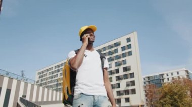 Yakışıklı Afro-Amerikan yemek dağıtım kuryesi kameranın önünde caddede poz veriyor. Sarı şapkalı ve sırtında ısı yalıtımlı çantasıyla telefonda konuşan gülümseyen bir adam.