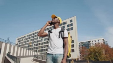 Yakışıklı bir Afro-Amerikan yemek kuryesinin portresi. Sokakta kameranın önünde poz veriyor, telefonla konuşuyor ve şehirde bir adres arıyor. Termal çantalı kurye.