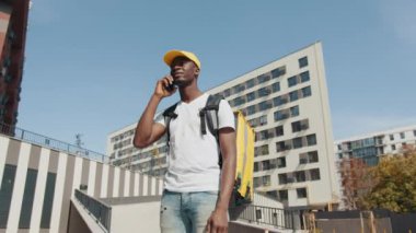 Sokakta kameranın önünde poz verip adres arayan yakışıklı genç yemek kuryesinin portresi. Afrika teslimatı, fast food siparişi vermek için cep telefonu kullanan bir müşteriyle konuşuyor..