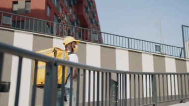 Teslimat konsepti. Tıbbi maskeli bir Afro-Amerikan kurye şehir sokaklarındaki bir müşteriye yemek paketi teslim ediyor. Kurye elinde telefon olan müşterinin adresini arıyor.