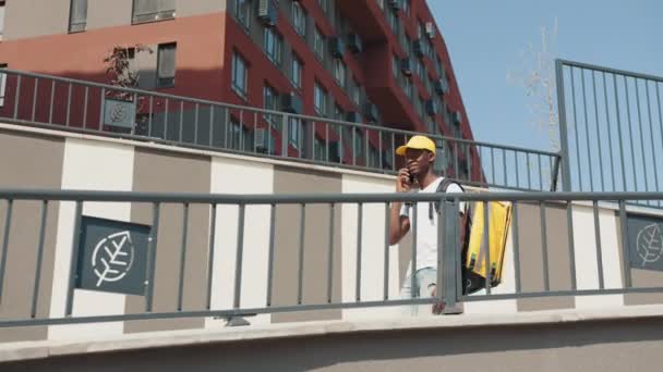 Un courrier afro-américain avec un sac à dos thermique parle au téléphone avec un client, descend la rue et livre des commandes aux clients. Journée ensoleillée dans une ville moderne avec des bâtiments élégants. — Video