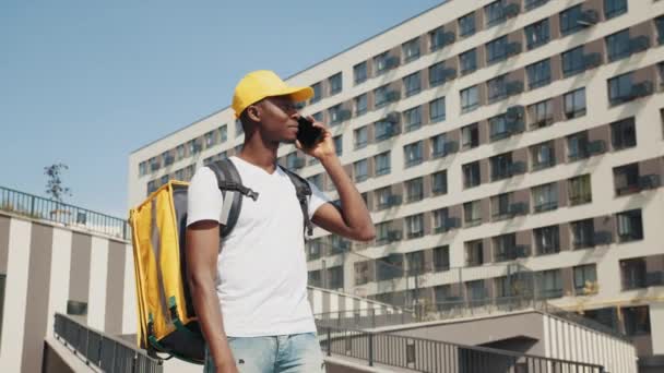 African delivery speaks to a customer using a mobile phone to order fast food delivery. Portrait of handsome young food delivery courier posing in front of camera on the street, looking for address — Stock Video