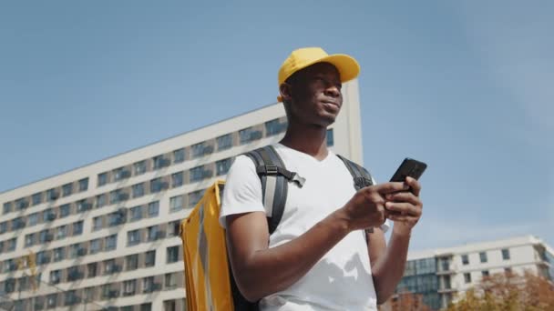Seorang kurir Afrika-Amerika memberikan makanan dengan ransel termal, berdiri di jalan, melihat navigasi pada smartphone. Karyawan pemasok memberikan pesanan kepada pelanggan secara online — Stok Video