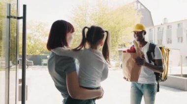 Afro-Amerikalı bir kurye, çocuğu olan güzel bir kadına, evin önündeki bir müşteriye yiyecek, meyve ve sebze paketi getiriyor. Ekspres gıda teslimatı ve online alışveriş kavramı