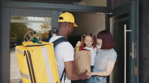 An African-American courier with a thermal backpack brings a package of food, fruit and vegetables to a beautiful woman with a child, a client in front of the house. Express delivery service — Stock Video