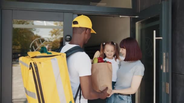 Termal sırt çantalı bir Afro-Amerikalı kurye, çocuklu, müşterisi olan güzel bir kadına yiyecek, meyve ve sebze paketi getiriyor. Ekspres dağıtım servisi — Stok video