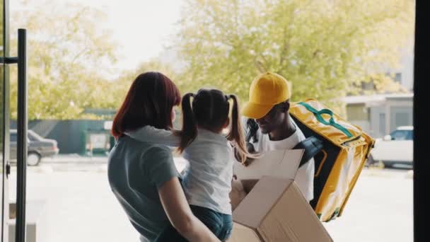 Il corriere porta un pacco per una bambina con sua madre alla porta di casa. Aprire il pacco, un bambino felice prende un peluche peluche giocattolo fuori dalla scatola. Acquista nei negozi online di e-commerce — Video Stock