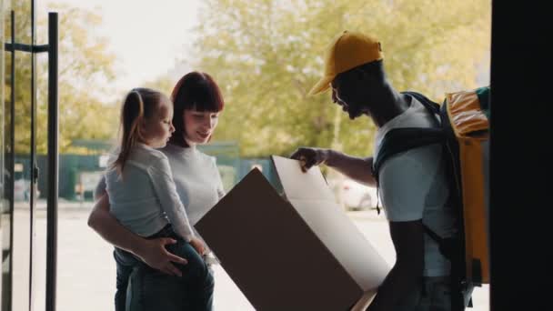 Der Kurier bringt ein Paket für das Mädchen und ihre Mutter an die Haustür. Beim Öffnen des Pakets holt ein glückliches Kind einen flauschigen Teddybär aus der Schachtel. Online-Shops im E-Commerce kaufen — Stockvideo