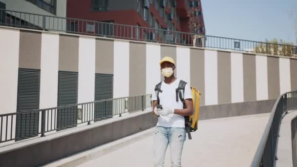 Hombre africano joven que trabaja en la entrega de alimentos y la celebración de teléfonos inteligentes. Precauciones de seguridad y entrega de alimentos durante un brote de coronavirus. El mensajero entrega comida rápida a la oficina o al hogar — Vídeos de Stock