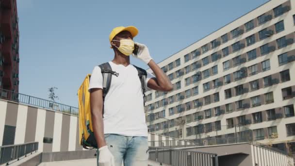 Retrato de un joven afroamericano con una máscara médica parado cerca de la casa en un día de verano y hablando por teléfono. Primer plano del mensajero alegre masculino. Entrega hombre con una sonrisa en la calle. — Vídeos de Stock