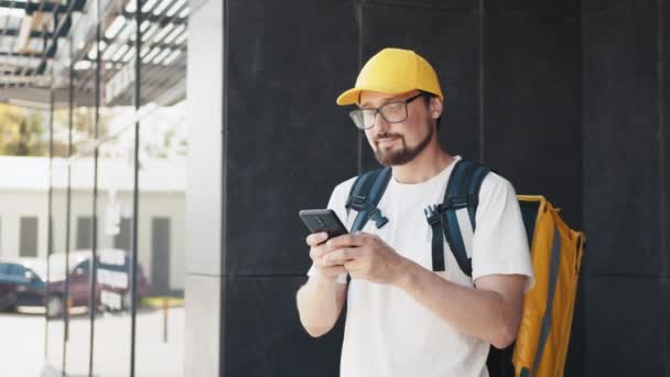 Happy delivery man writes text messages on his smartphone. On his shoulders and cap he wears a yellow backpack for delivery. The food vendor controls the phone in the city — Stock Video