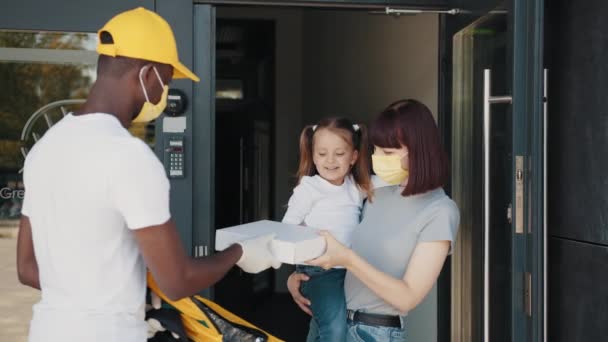 Un mensajero con guantes y una máscara entrega comida y bienes durante una pandemia de coronavirus. Entrega de compras en línea. Tecnologías durante la pandemia del coronavirus. Mamá e hija reciben un paquete — Vídeos de Stock