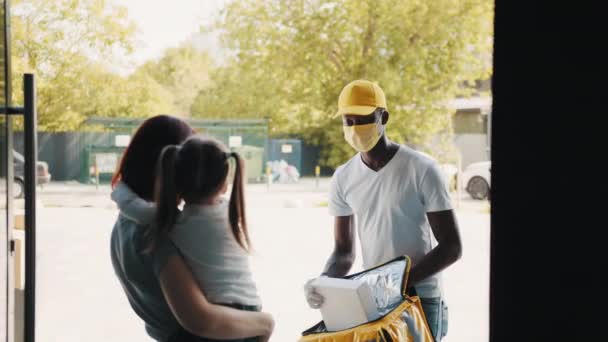 Eldiven ve maske takmış bir Afro-Amerikalı kurye, koronavirüs salgını sırasında yemek ve yiyecek dağıtıyor. Anne ve kızı bir paket alıyorlar. Çevrimiçi mağazalar. Koronavirüs salgını sırasındaki teknolojiler. — Stok video