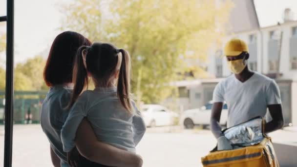 Portret van een Afro-Amerikaanse koerier in handschoenen en maskers die voedsel en goederen leveren tijdens een coronaviruspandemie. Mam en dochter krijgen een doos. Online winkels. Levering tijdens een coronaviruspandemie. — Stockvideo