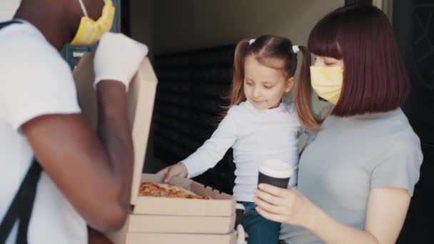 Красивая молодая улыбающаяся женщина с ребенком у двери своего дома и встречает доставщика пиццы, который дает ей картонные коробки, полные вкусной приготовленной пиццы. Семья из двух человек с нетерпением едят пиццу — стоковое видео