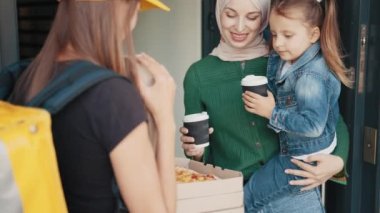 Bir kurye kız kutularca pizza ve içecek getiriyor, müslüman bir kadına ve çocuğuna evinin kapısında gülümsüyor, pizza gösteriyor. Cafe ve restoran Teslimat Servisi. Yemek teslimatı.