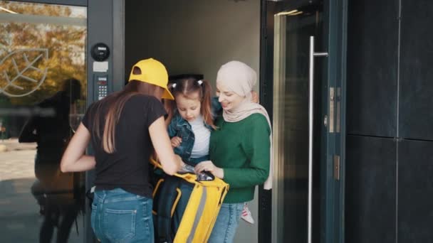 Una mujer musulmana positiva en un hiyab con un niño acepta su pedido en línea de una mensajera con gorra amarilla y mochila térmica. Concepto de entrega. El mensajero sostiene y da una caja de cartón — Vídeos de Stock