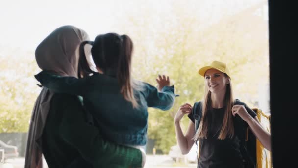 Una joven mensajera trajo un paquete a la familia. Una mujer musulmana positiva en un hiyab con un niño acepta su pedido en línea de una mensajera con gorra amarilla y mochila térmica. Concepto de entrega — Vídeos de Stock