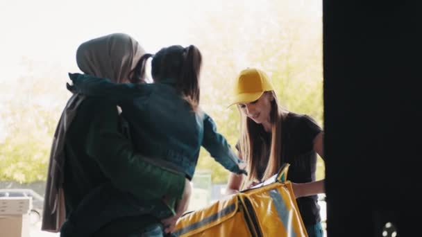 Szállítási koncepció. Egy fiatal futár lány hozott egy csomagot a családnak. Egy pozitív muszlim nő egy hidzsábban egy gyerekkel elfogadja az online rendelését egy sárga sapkás és termikus hátizsákos futártól. — Stock videók