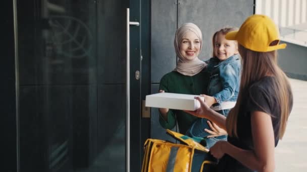 Paczka dla dziecka. Kurierka przyniosła paczkę rodzinie. Pozytywna muzułmanka w hidżabie z dzieckiem przyjmuje zamówienie online od kurierki w żółtej czapce i termalnym plecaku — Wideo stockowe