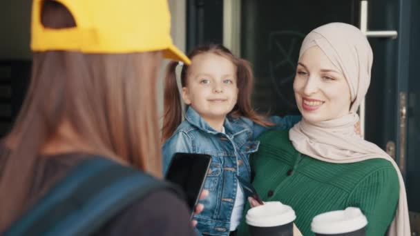 Entrega de comida rápida y pago sin contacto sin efectivo. Feliz mensajera con sombrero amarillo sostiene un teléfono y cajas de pizza, una mujer musulmana en un hiyab con un niño paga con tarjeta de crédito — Vídeos de Stock