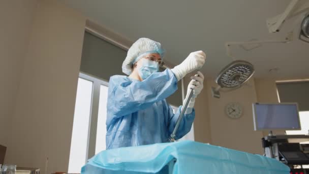 Πορτρέτο μιας εργαζόμενης στον τομέα της υγείας με προστατευτική μάσκα που στέκεται στο τραπέζι με χειρουργικά εργαλεία. Χειρουργικό όργανο. Χειρούργος κρατά ένα όργανο για χειρουργική επέμβαση. Προετοιμασία για λαπαροσκόπηση — Αρχείο Βίντεο