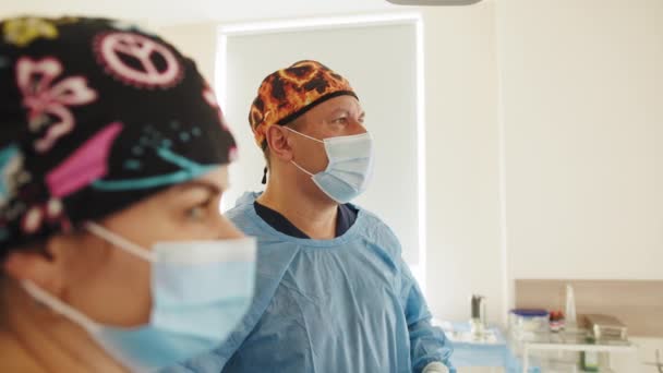 Surgeon performing operation in operating room. Caucasian surgeon male healthcare professional in a hospital operating theatre wearing a surgical cap and mask. Foreground nurse — Stock Video