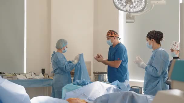 Kirurgi, medisin og pasientbegrep - sykepleier assisterer kirurg og hjelper til med å beskytte slitasje før operasjon i operasjonsrommet på sykehuset – stockvideo