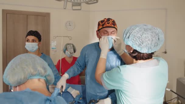 Zespół chirurgiczny pracujący w Operacyjnym Teatrze. Podczas operacji na sali operacyjnej pielęgniarka lub asystentka przytwierdza maskę do zmęczonego lekarza. Skoncentrowany zespół chirurgiczny w sali operacyjnej. — Wideo stockowe