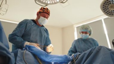Asistanlarıyla birlikte düşük açılı cerrah higroma ameliyatından sonra düzgün dikişlerle karın derisine dikiş atıyor. Doktor, hastanedeki ameliyathanede yaraya dikiş dikiyor.