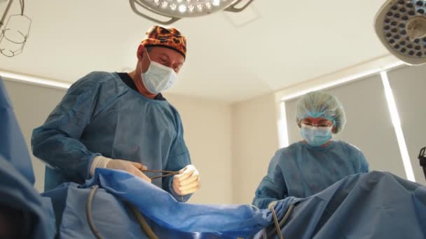 Låg vinkel av koncentrerad kirurg man med sina assistenter suturer magen hud under operation med snyggt stygn efter avlägsnande hygroma. Läkare syr på sår i operationssalen på sjukhus — Stockvideo