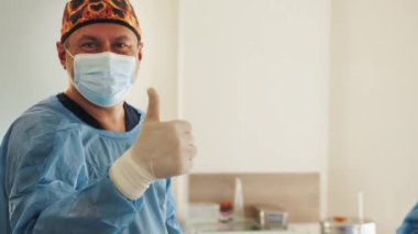 Başarılı bir operasyondan sonra gülümseyen ve kameraya bakan profesyonel cerrahın portresi. Arka plan Modern Hastane Ameliyat Odası 'nda. Gülümseyen doktor başparmağını kaldırıyor.