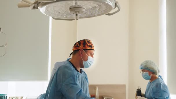 Portrait du chirurgien professionnel regardant dans la caméra et souriant après une opération réussie. En arrière-plan Salle d'opération moderne de l'hôpital. Le médecin éteint la lampe chirurgicale — Video