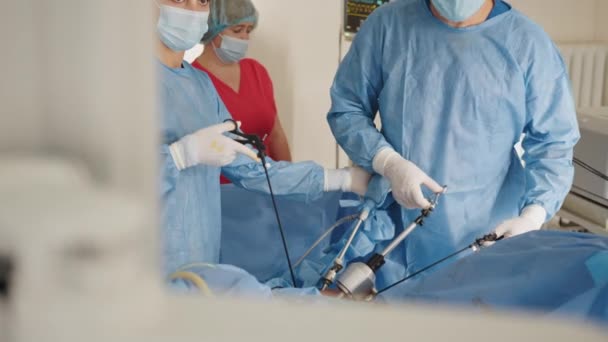 Портрет женщины-медработницы в защитной маске и стерильных перчатках, держащей лапароскопический захват с троакаром, стоя рядом со столом с хирургическими инструментами. Хирург и его ассистент размыты — стоковое видео