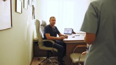 Klinikte masasında oturan erkek doktor, kadın hastanın tıbbi geçmişini okuyor ve muayene sırasında onunla konuşuyor. Aile Doktoru Hastaya Teşhisi Açıklıyor. Aile Sağlığı