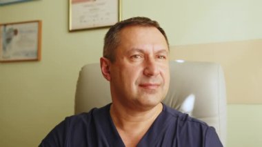 Tıp bürosunda oturan kıdemli doktorun portresi. Gülümseyen beyaz erkek doktor portresi hastane masasında oturmuş kameraya bakıyor. Kıdemli doktor ya da terapist, sağlık hizmeti konsepti