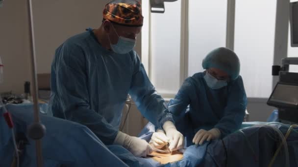 Команда хірургів зшиває тіло пацієнтів. Тримач голки та хірургічна нитка в руках хірурга. Лікарі носять стерильні рукавички, хірургічну форму та хірургічні маски. шви шкіри — стокове відео