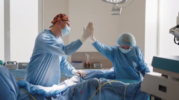 Erkek ve kadın cerrah hastanede birbirlerine beşlik çakıyor. İki yakışıklı doktor beşlik çakıyor ve gülümsüyor. Modern bir ameliyathanede duruyorlar. Hastanede ameliyattan sonra cerrah. — Stok video